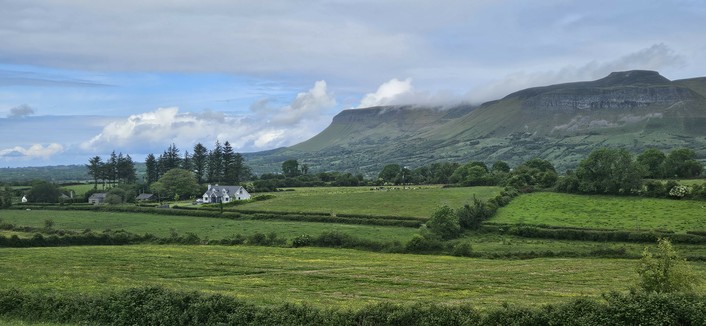 A lush green landscape features a patchwork of fields, hedgerows, and a few white houses nestled among trees. In the background, a dramatic mountain ridge rises, partly shrouded in low cloud, under a blue sky with scattered clouds.