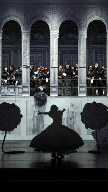 Silhouette of a woman twirling around in the middle of a stage with her arms raised, large painted flowers to her left and right. The stage set in the background is white with black line drawings: a gallery with high arcades. The opera chorus stands on the gallery between the arcades, dressed in black and holding small white flags, gazing happily at the woman.  Soprano Megan Marie Hart as Elvire in Paul Georg Dittrich's staging of Daniel Auber's grand opéra La Muette de Portici at Staatstheater Darmstadt, 2025. Photo: © Benjamin Weber / Staatstheater Darmstadt
