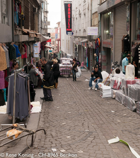 Das Bild zeigt eine schmale Kopfsteinpflasterstraße in einem belebten Marktgebiet in Istanbul. Auf der linken Seite sind verschiedene Kleidungsstücke auf Ständern und an Markisen ausgestellt, und die Menschen stöbern und unterhalten sich mit den Verkäufern. Auf einem Schild steht "ÖRÜCÜLÜ CARŞISI" und "TEXTIL CENTER", was auf einen Textilmarkt hinweist. 
Auf der rechten Seite befinden sich weitere Geschäfte mit geschlossenen Fensterläden, und man sieht einige Menschen auf dem Boden sitzen, die möglicherweise eine Pause machen oder warten. es handelt sich hier um sogenannte Lastenschlepper welche in diesem Stadtviertel einen Grossteil des Transportes in den Gassen übernehmen
