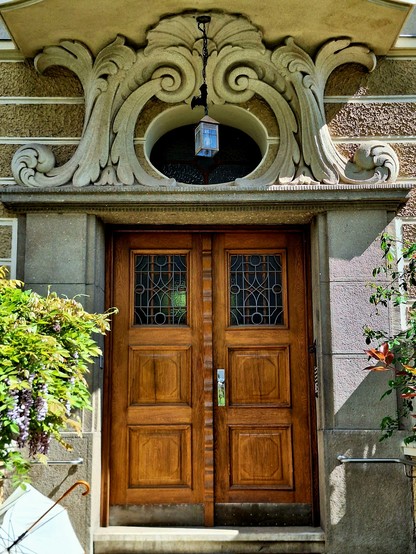 Picture of a wooden entrance door. Over the door there is a small oval window, surrounded by floral decorations. These decorations are symmetric and also seem to form two eyes, making the window look like an open mouth. In front of the window, a small lantern with an iron bat is hanging from the balcony above.