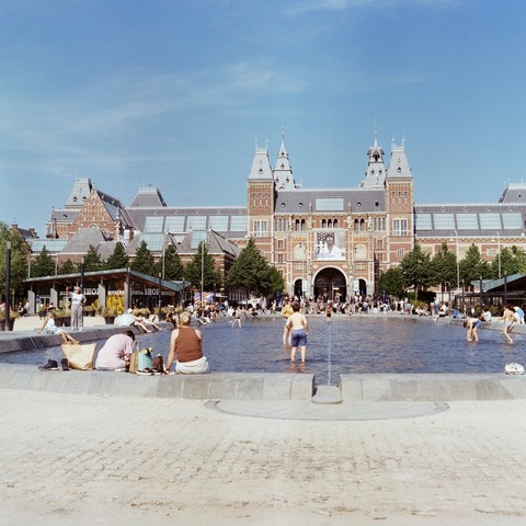 Scan of analoge square photograph of Amsterdam Museumplein, with in background Rijksmuseum, with a poster of American Photography exhibition over main tunnel entrance and in foreground people playing in shallow pond or sitting on the concreet sides of the pond. Hot day at start of May 2025.