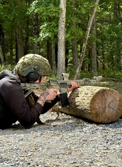 Das Foto zeigt eine Person mit einem Goat Tattoo iauf dem rechten Handgelenk n einem schwarzen Hoddie mit Helm und Gehörschutz, die hinter einem Baumstamm liegt, mit einer angeschlagenen Colt AR-15 ein nicht sichtbares Ziel im Visier.