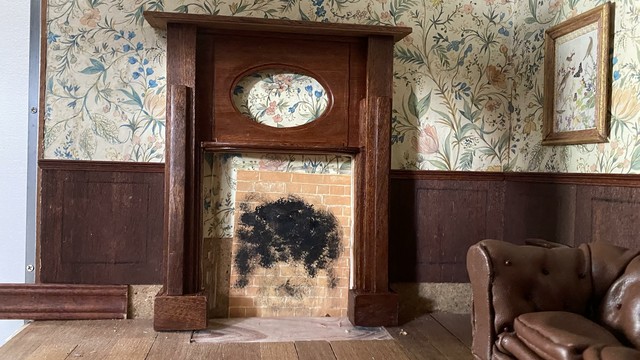 A sitting room in a dolls house with a chesterfield sofa pushed against the right wall, light pours in from the open door to the left. Bits of the skirting are loosely arranged against the wall. There’s dark wood paneling and a floral wallpaper. The floor is boarded with hand cut boards.