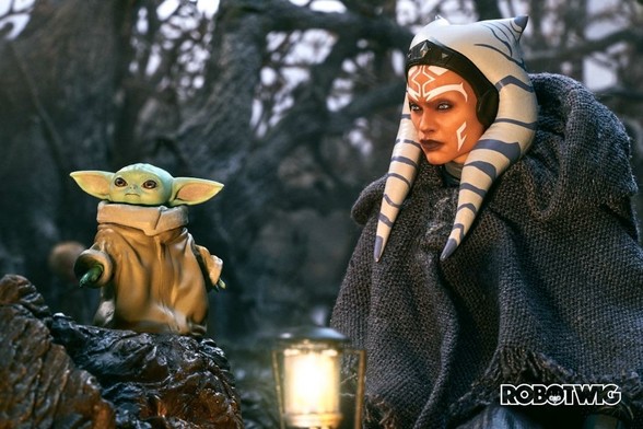 Ahsoka and Grogu sit by a glowing lamp deep in the woodland. Grogu looks up to the sky. Ahsoka looks at him
