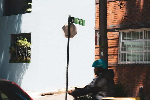 Street scene with a blurred person on a bicycle in the foreground wearing a blue helmet. A street sign for "Pine" is mounted on a pole next to a stop sign. The background features a light blue building with a window and plants, adjacent to a red brick building.