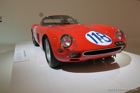 The image features a classic red sports car displayed in a museum setting. The car is a Ferrari 250 GTO, identifiable by its distinctive design and the number "118" on the front. The car has a sleek, aerodynamic shape with a low profile, round headlights, and a prominent front grille. The body is painted in a vibrant red color, and the car is positioned on a white platform, which enhances its visual appeal. The car's design includes side vents and a streamlined shape, typical of high-performance vehicles from the era. The background is minimalistic, with a plain wall and a small informational plaque next to the car, providing context about the vehicle. The lighting is focused on the car, highlighting its curves and details. The image is taken from a slightly elevated angle, showcasing the car's front and side profile.

―

The image features a close-up view of a red car's rear wheel. The wheel is equipped with a wire-spoke design, which is characteristic of classic sports cars. The wire spokes are silver and radiate from a central hubcap, which is also silver and features a small emblem. The tire is black with visible tread and has the brand name "Dunlop" embossed on its sidewall. The car's body is a vibrant red, and the wheel is mounted on a white platform, suggesting the car is on display. The background is blurred, focusing attention on the wheel and tire. The image is taken from a low angle, emphasizing the wheel's details.