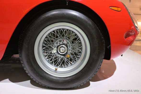 The image features a close-up view of a red car's rear wheel. The wheel is equipped with a wire-spoke design, which is characteristic of classic sports cars. The wire spokes are silver and radiate from a central hubcap, which is also silver and features a small emblem. The tire is black with visible tread and has the brand name "Dunlop" embossed on its sidewall. The car's body is a vibrant red, and the wheel is mounted on a white platform, suggesting the car is on display. The background is blurred, focusing attention on the wheel and tire. The image is taken from a low angle, emphasizing the wheel's details. The text "Nikon Z 3, 40mm, f/5.6, 1/60s