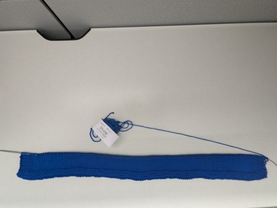 The edge of a blanket worked on a chiaogoo red lace circular needle.  The yarn is a navy blue and a deflated skein leads into one corner of the blanket.  There is a yarn wrapper indicating the yarn is Knit Picks Shine Worsted in color French Blue.