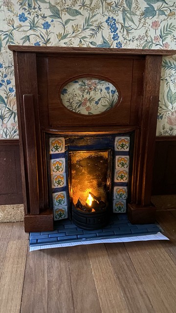 A fire burning cozily in a dolls house grate with a fire surround propped up around it. Nothing is glued and you can see the paper under the hearth but it is super nearly there. The wall paper is a floral one and there’s dark wood panelling on the walls.