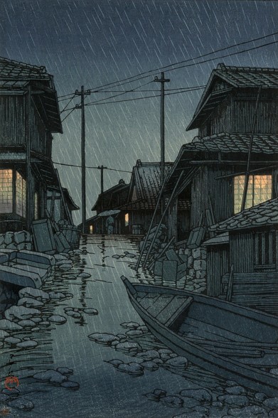 Rain falls on a narrow lane in a town by the seaside in Kawarago, Miyagi Prefecture. In the foreground, a small wooden boat is pulled up, lying at an awkward angle on the uneven, stony ground. Beyond, warm lights glow in the windows of houses on either side of a meandering little lane; up ahead, a single pedestrian carrying an umbrella passes beneath wires strung between utility poles. Rain continues to fall across the scene; the ground is slick with rainwater, and the sky above is dark and overcast.