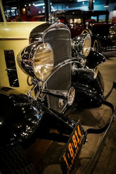 he image features a close-up view of the front of a vintage car, likely from the 1930s, displayed in a museum setting. The car's front grille is prominent, with a vertical chrome bar design and a large, round headlight on each side, reflecting the ambient light. The car's body is painted in a glossy black finish, with a contrasting cream-colored section visible on the left side. The license plate reads "1-814 COLORADO 1932," indicating the car's registration year and state. The car's hood ornament is visible at the top center of the grille, adding to its classic aesthetic. The background shows other vintage cars, suggesting a collection or exhibit. The lighting is warm, highlighting the car's polished surfaces and intricate details.

Provided by @altbot, generated privately and locally using Ovis2-8B