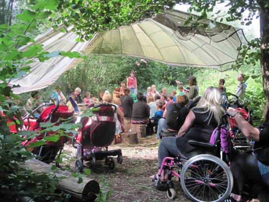 Community gathering outdoor under a large canopy, people listening to a man playing a guitar.
