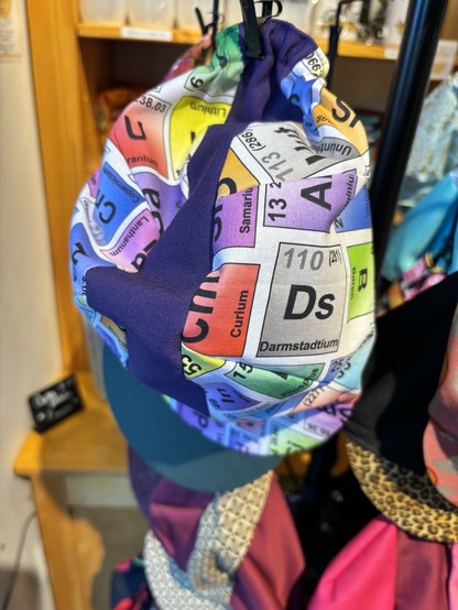 A colorful fabric cycling cap featuring a design inspired by the periodic table of elements, showcasing various chemical symbols and atomic numbers. The bag is displayed on a rack in a store.