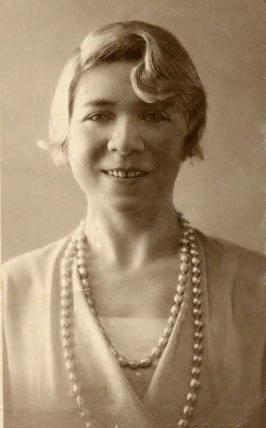 Retrato sepia de una mujer joven, Alfonsina Storni, con el pelo corto y ondulado, peinado hacia atrás de la cara. Lleva un vestido claro y un collar de perlas de doble hilera. Su expresión es seria pero amable, y su mirada se dirige directamente al espectador. La imagen tiene un estilo fotográfico antiguo.