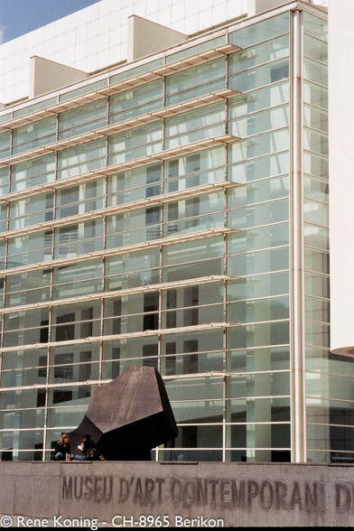 The image shows a modern building with a glass facade, featuring a series of horizontal metal beams that create a grid-like pattern. The building is part of the Museu d'Art Contemporani de Barcelona, as indicated by the text on the stone wall in the foreground. 
The structure is predominantly made of glass, with reflections visible on the surface, and the upper part of the building is white. In the foreground, there is a large, dark abstract sculpture, and two people are sitting on a ledge in front of the building, one of whom is holding a drink. The sky is clear, suggesting a sunny day.