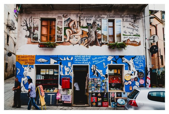Auf dem Foto ist eine farbenfrohe Wand mit auffälliger Straßenkunst zu sehen, die verschiedene Szenen und Figuren darstellt. Die Wand ist überwiegend mit kräftigen blauen und erdigen Tönen gestaltet, während die Fenster mit hölzernen Rahmen und Blumenkästen den Eindruck von Charme und einladender Atmosphäre vermitteln. Vor der Wand stehen Passanten, die neugierig die ausgebreitete Kunst und die Auslagen in einem kleinen Laden betrachten. Dieser Ort strahlt eine lebendige Kreativität aus und lädt dazu ein, die Geschichten und Geheimnisse hinter den Kunstwerken und der Umgebung zu entdecken.
