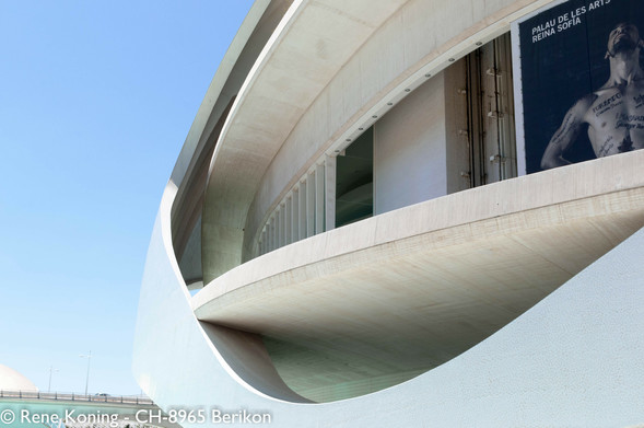 The image features a modern architectural structure, likely a museum or cultural center, with a distinctive curved design. The building's facade is predominantly white, with smooth, flowing lines that create a sense of movement and fluidity. The structure is composed of concrete, with a large, sweeping curve that forms a prominent part of the building's exterior. The upper section of the building has a large, dark poster with the text "PALAU DE LES ARTS REINA SOFIA" and an image of a shirtless man with tattoos. The sky is clear and blue, indicating a sunny day. The building's design is contemporary, with a minimalist aesthetic that emphasizes form and function. The image captures the building from a low angle, highlighting its impressive scale and architectural features. It is situated in Spain, city of Valencia