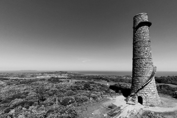 A striking black and white photograph of the tall, cylindrical stone chimney of Ballycorus Lead Mines, also known as Carrickgollogan Tower, standing prominently on a hilltop. A spiral staircase wraps around its exterior. In the background, a vast landscape extends towards the sea under a clear sky, with a town visible in the distance. The foreground shows rugged terrain and low-lying vegetation.