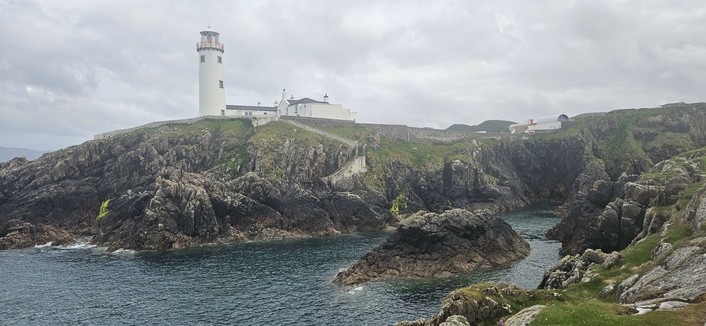 A white lighthouse stands atop rugged rocky cliffs overlooking a small bay. The sea below is deep blue, and the sky is overcast, adding to the wild, remote feel of the coastal landscape