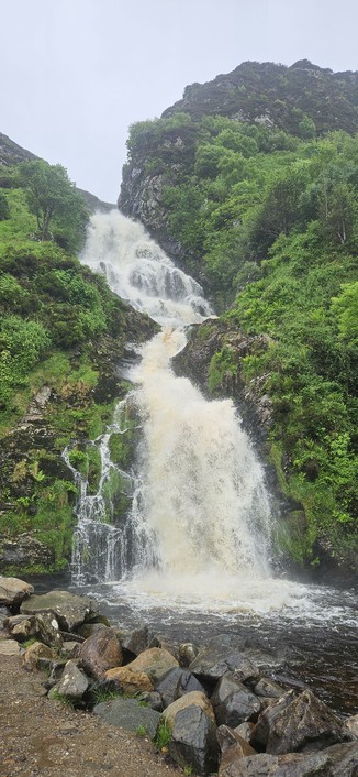A powerful waterfall cascades down a rocky, green hillside, surrounded by dense vegetation. The water crashes into a pool at the base, with large rocks and boulders scattered along the edge.