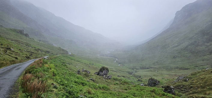 A misty mountain valley with rolling green slopes and tufts of brown grass in the foreground. The distant hills are barely visible through the dense fog, creating a tranquil and atmospheric landscape.