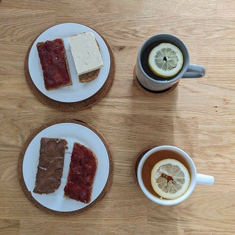 Auf dem Bild ist ein schlicht angerichtetes Frühstück für zwei Personen auf einem hellen Holztisch zu sehen.
Linke Seite:

    Zwei kleine Teller mit je zwei Brotscheiben:

        Oben: eine Scheibe mit roter Marmelade, die andere mit Tofu belegt.

        Unten: eine Hälfte mit Schokocreme (vermutlich Nuss-Nougat) und die andere ebenfalls mit roter Marmelade.

Rechte Seite:

    Zwei Tassen mit Tee, jeweils mit einer Zitronenscheibe oben schwimmend:

        Oben: eine graue Tasse

        Unten: eine weiße Tasse

Beide Teller und Tassen stehen auf runden Korkuntersetzern. Die Gesamtkomposition ist einfach, aber liebevoll – ein typisches veganes oder vegetarisches Frühstück mit süßen und herzhaften Komponenten.