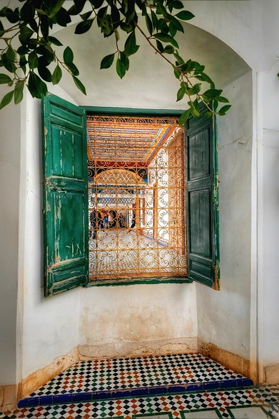A vibrant green wooden window with open shutters in a white stucco wall. The window is adorned with intricate golden wrought ironwork, revealing a colourful, patterned interior courtyard with arches and columns. The floor beneath the window is tiled with a black and white checkerboard pattern, bordered by colourful geometric tiles. Lush green leaves from an unseen plant gracefully hang into the top left of the frame.