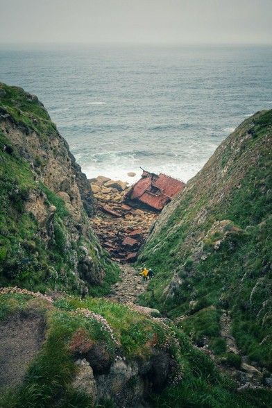 Looking down a gully between two steep rock walls, towards a beach and the sea. On the narrow section of beach visible at the end is a rusted hull of a ship. Two small figures can be made out walking along the gully.