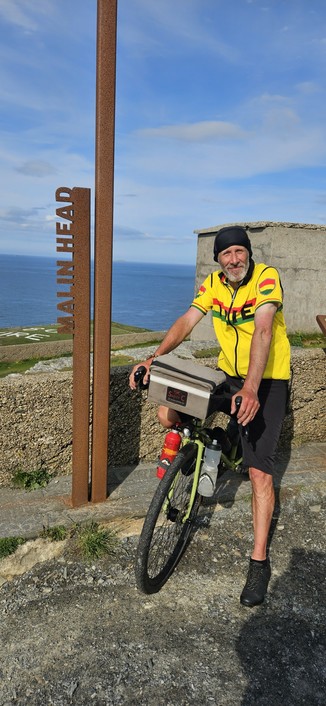 A cyclist wearing a yellow, red, and green jersey stands with their touring bicycle at Malin Head, Ireland's northernmost point. The cyclist is next to a tall, rusted metal sign that reads "MALIN HEAD," with the Atlantic Ocean and a partly cloudy sky in the background. The bicycle is equipped for long-distance riding, featuring a handlebar bag, water bottles, and bikepacking gear. The scene conveys a sense of achievement and adventure at a scenic coastal landmark.