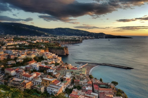 A panoramic shot of Sorrento, Italy, at sunset. The town's colourful buildings cascade down a cliff face towards the calm blue sea. A long pier extends into the water, and in the distance, hills recede under a dramatic sky with both clear patches and dark clouds, illuminated by the golden light of the setting sun.