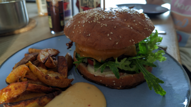 ein großer Burger aus dem etwas Rucola herausragt, Kartoffelecken und Chilicheesesauce auf einem blaugrauen Teller
