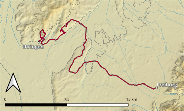 Das Bild zeigt eine topografische Karte, die eine Route von Ihringen nach Freiburg darstellt. Die Route ist in Rot markiert und verläuft durch eine hügelige Landschaft. Die Karte zeigt die Höhenlinien, die die topographische Struktur der Region veranschaulichen. Die Route beginnt in Ihringen im Nordwesten und führt nach Süden bis nach Freiburg im Südosten. Ein Kompass in der linken unteren Ecke zeigt die Richtung Nord. Eine Skala am unteren Rand der Karte zeigt die Entfernung in Kilometern, wobei 7,5 km und 15 km markiert sind. Die Karte ist in einem hellen Beige-Ton gehalten, mit blauen Linien, die Flüsse oder Gewässer darstellen.