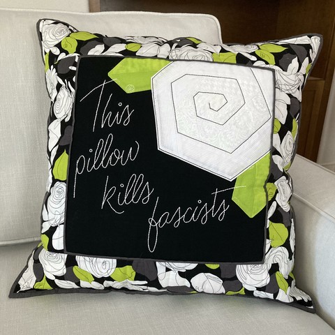 Large quilted pillow displayed on the corner of a white couch. “This pillow kills fascists” is hand embroidered in a white script font on the black center of the pillow. A wonkily pieced white rose, nestled among two lime green wonkily pieced leaves, fills the upper right corner; it is outline stitched in a thick black embroidery thread to echo the border fabric. The border fabric features large white scribble roses with grey and lime leaves on a black background. It’s one of my favorite fabrics, and I was unwilling to cut into it for the longest time, but it turned out to be perfect for this anti-fascist craftivism project.