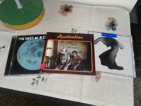 An inage showing a table with three music CDs. From left to right, the CDs include "The Best of R.E.M. 1988–2003” by R.E.M., “Here Comes the stunts” by AWOLNATION, and “Fragrant World” by Yeasayer. The R.E.M. and Yeasayer discs are in jewel cases, while the AWOLNATION disc is in a paper sleeve.
