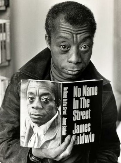 Black and white photo of author James Baldwin holding a copy of his book "No Name In The Street."