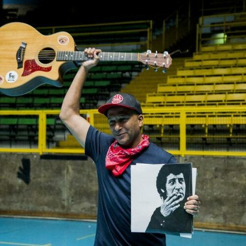 Tom Morello con gorra roja y pañuelo rojo al cuello, sostiene una guitarra acústica sobre su cabeza con su mano izquierda. Con su mano derecha sostiene una imagen en blanco y negro de Victor Jara fumando. El fondo es una cancha deportiva con gradas amarillas.