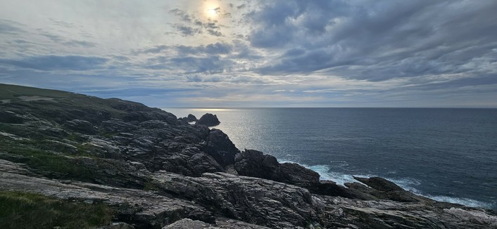 A dramatic rocky coastline under a mostly cloudy sky, with the sun partially visible behind the clouds. The sea is calm, reflecting the light from the sky, and rugged cliffs dominate the foreground.