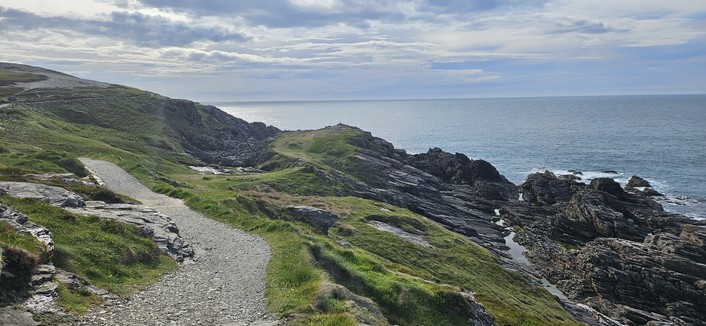 A winding gravel path runs along a grassy, rocky headland overlooking the ocean. The coastline is rugged with jagged rocks leading down to the water, and the sky is partly cloudy with soft light.
