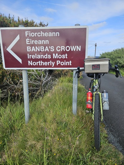 A bilingual road sign in Irish and English reads, "Fiorcheann Éireann BANBA'S CROWN Irelands Most Northerly Point," with an arrow pointing left. A touring bicycle with water bottles and a handlebar bag is parked next to the sign, and green grass surrounds the area.