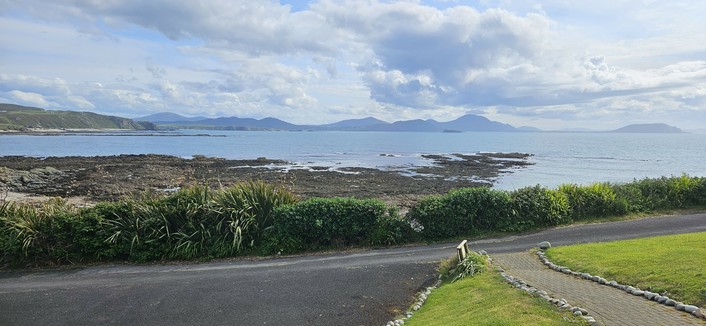 A tranquil coastal scene with a rocky shoreline and calm sea. In the distance, rolling hills and mountains rise under a partly cloudy sky. The foreground includes a neatly maintained lawn and a paved path bordered by low shrubs.