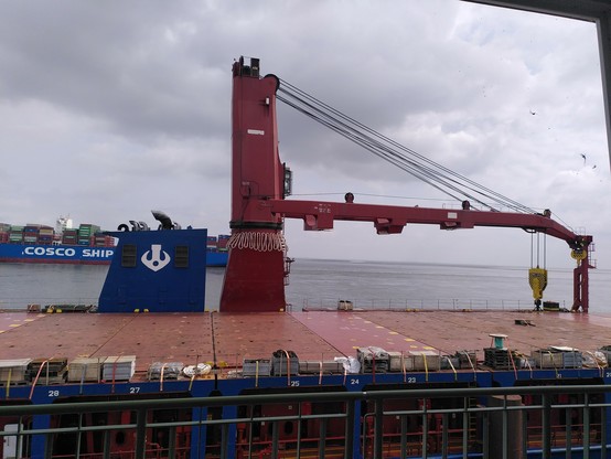 Schiffskran, um den ein dickes Tau in Buschten hängt. Im Hintergrund ein Großcontainerschiff