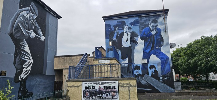 A large mural painted on the side of a building shows several figures in suits running, with one figure in the foreground wearing a blue suit. Another mural on the adjacent wall depicts a person in dark clothing holding a rifle. Below, a sign references the INLA (Irish National Liberation Army) and ICA, with slogans about allegiance and resistance. The murals are painted in shades of blue, black, and white, and the scene is set in a residential area with a cloudy sky above