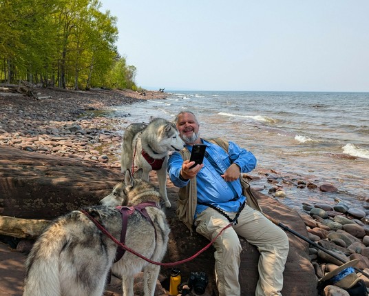 Jeff sits on a large emerging bit of shale bedrock poking out of an otherwise rocky shore of Lake Superior. He and Zeena are both looking at his phone, either commenting on photos Jeff has taken or taking a selfie. One can't tell. Tonttu stands in the foreground, looking a bit bored. Lake Superior is calm, with a few white caps on waves just before hitting shore. The sky is a bit hazy from Ontario wildfire smoke blowing across the lake.