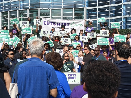 A crowd of people at a rally, listening to a Black woman with a microphone wearing a green shirt saying "United Domestic Workers", backed by a multiethnic group of people holding up signs and banners that say "Cuts Hurt Care".