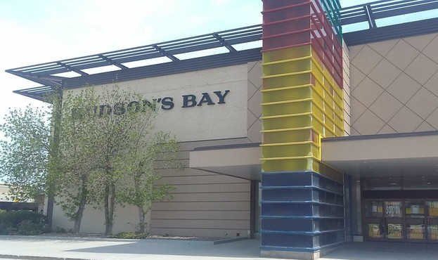 Colour photo of the St. James Street entrance of The Hudson's Bay Company store at Polo Park, in it's last design, which includes a three storey tall rectangular column dressed in The Bay's signature green, red, yellow, and indigo stripes, which are each several meters tall.
The entrance doors under the canopy are covered with "Store Closing" and 50% / 70% discount signage.
There are three youngish trees growing in front of the text logo.
The sky above shows high thin cirrus cloud cover, yet it is sunny all around.