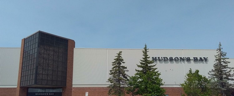 Colour photo of the Meadowood Entrance to the St. Vital Hudson's Bay Company store, which mostly evokes their latest "livery", yet the red brick and black pointed lattice structure above the doors is of the earlier 1970s/1980s "The Bay" design.
There are four trees visible below the store's text logo, which are situated to the right.
The sky above is a clear light blue.