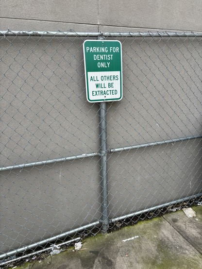 Grey wall with chain link fence directly in front of it. A green and white sign says “Parking for Dentist Only - All Others Will Be Extracted”. Some mossy cement is seen at the base of the fence.