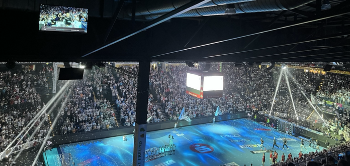 Foto von der THW Halle in Kiel. Voll besetzte Ränge, in der Mitte hängt ein Videowürfel an der Decke. Die Spieler laufen gerade ein. Der Boden ist blau ausgeleuchtet