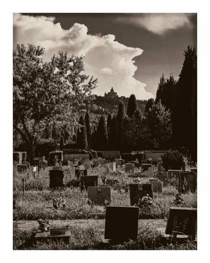 The photo is of cemetery with headstones of various shapes and sizes - with grass and scattered flowers around them. In the background there is a row of manicured cypress trees which helps lead your eye toward a hilltop with a silhouetted church (San Luca) which is in the mid ground of the photo. Above the hill in the background, and takes up roughly the too third of the photo, there are dramatic clouds in the sky.