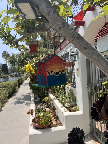 A small decorated birdhouse with the word “Venice” on it hangs on a tree branch.